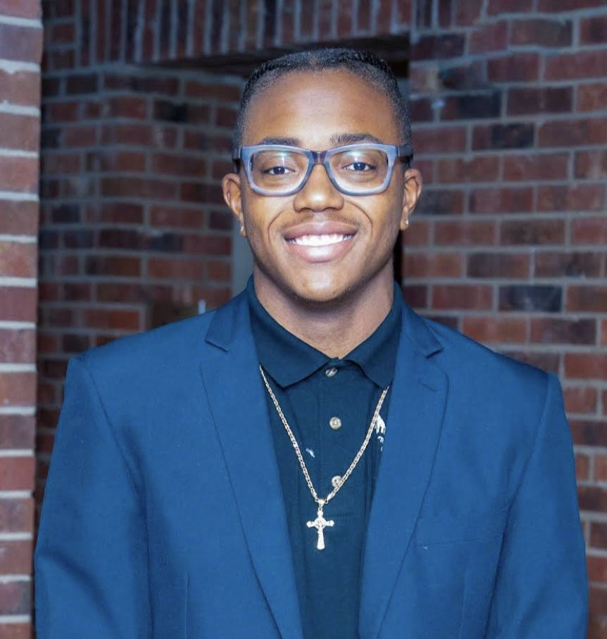 A young man wearing a blue suit poses for a photo in front of a brick wall.