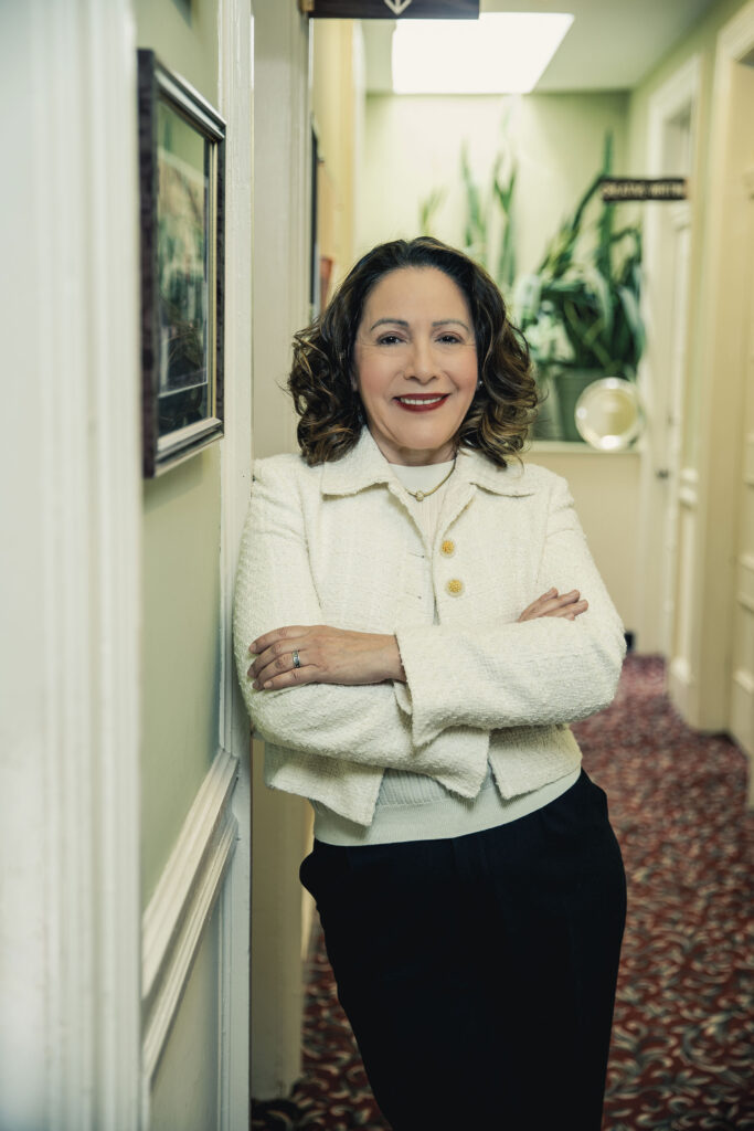 A woman leans against the hallway near academic offices. She wears bright red lipstick, business dress and her arms are folded across her chest.