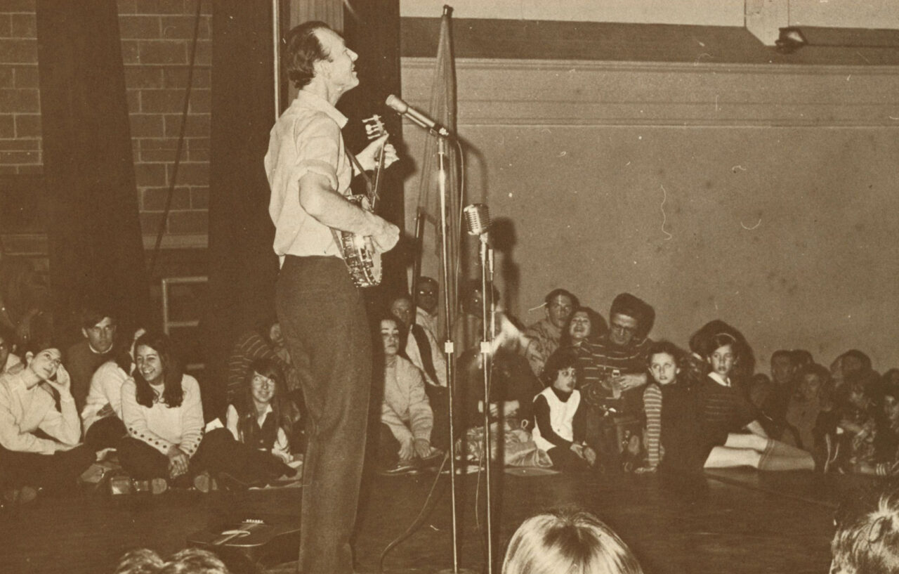 A folk singer strums a banjo and sings into a standing microphone as a sitting crowd watches. It's a vintage photo.