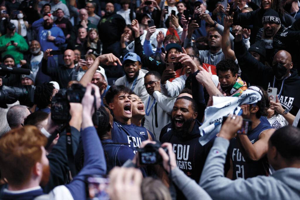 A crowd of sports fans cheers wildly. Some are wearing FDU shirts.