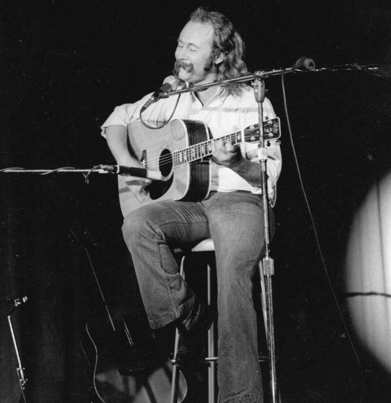 A male musician with long hair sits on a stool on stage, strumming an acoustic guitar and singing into a standing microphone.