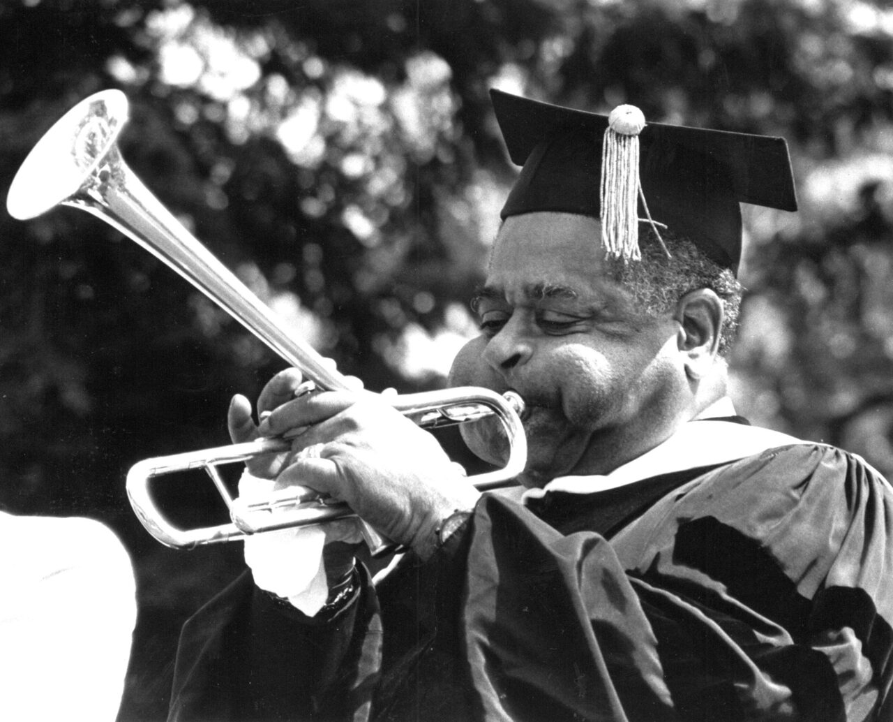 A man wearing a graduation cap and gown plays the trumpet.