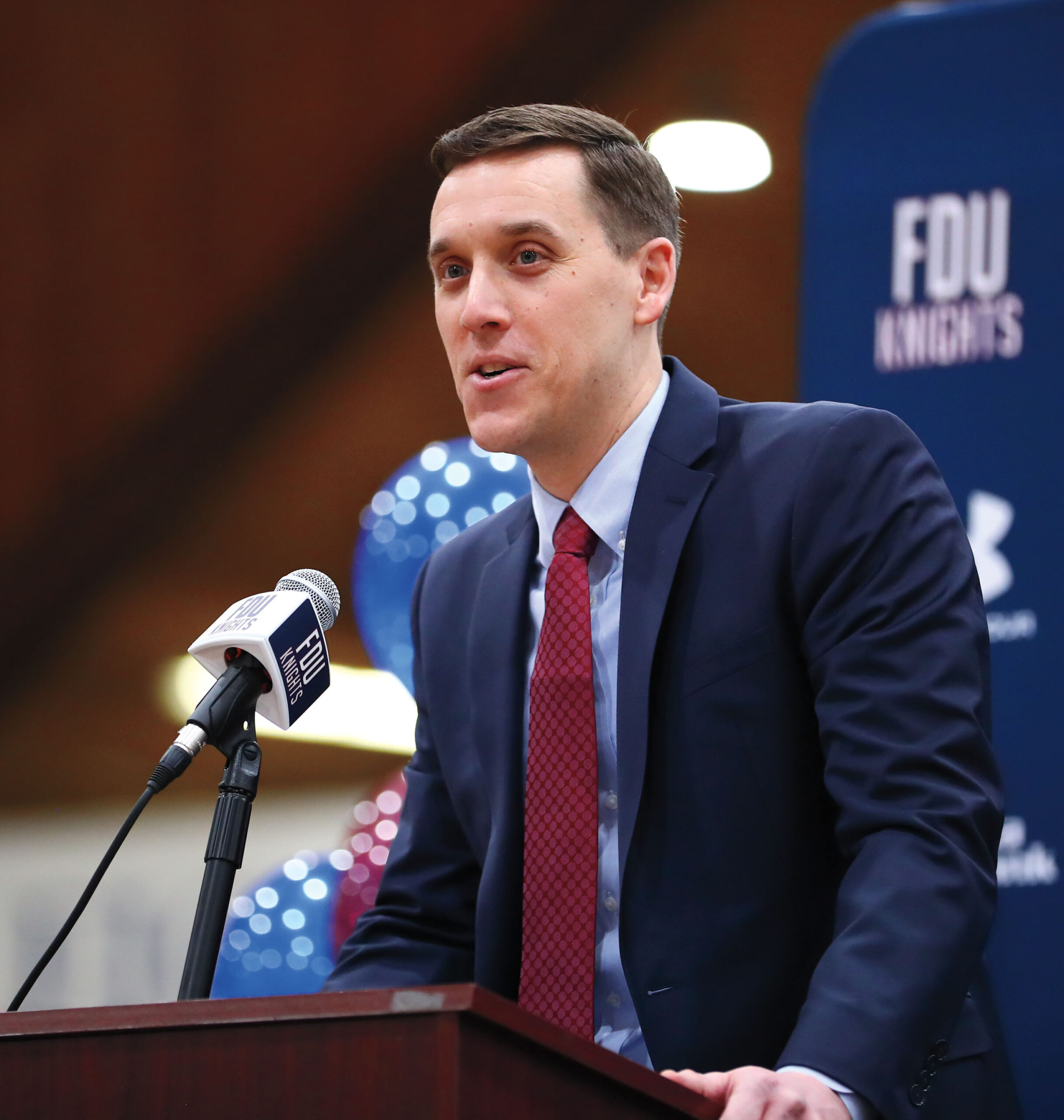 A man in a jacket and tie stands at a podium during a press conference.