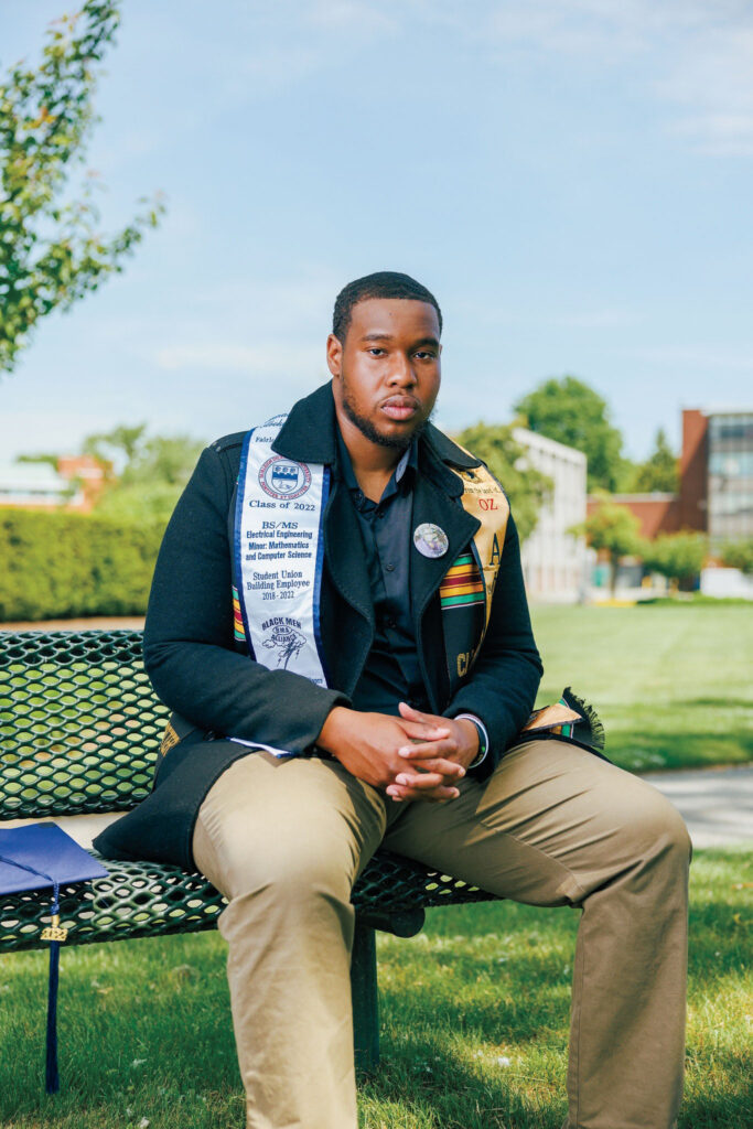 A young man wearing graduation stoles and accolades sits on a bench.