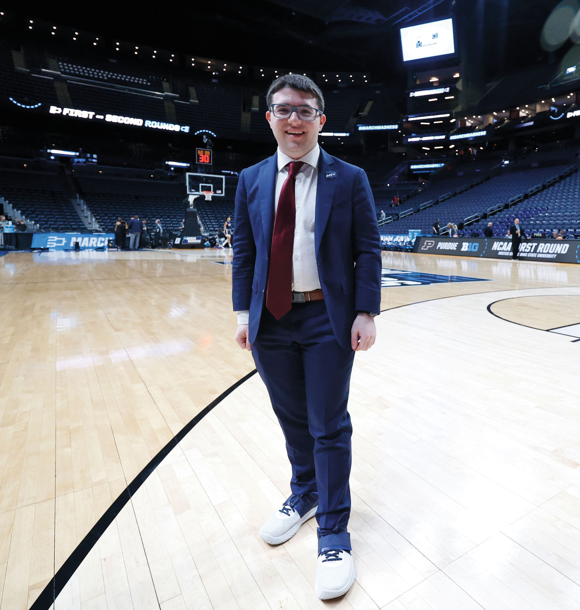 A young man in glasses, a suit and tie, stands in the middle of an empty basketball court in an arena.