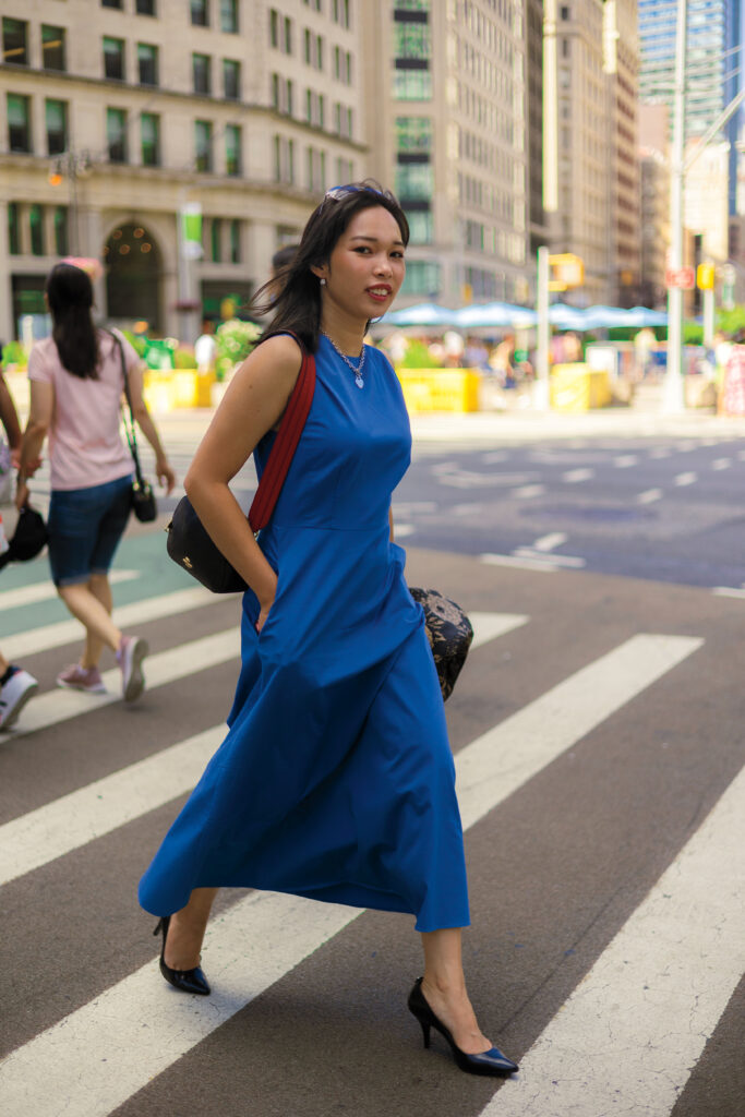 A young woman wearing a bright blue dress crosses a crosswalk in New York City.