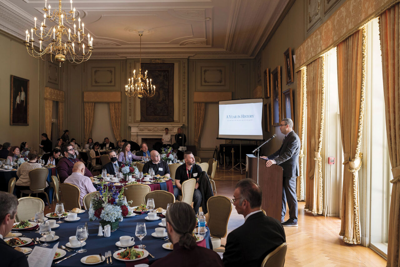 A banquet in a ballroom, with a lot of people sitting at tables to eat. A man in a suit stands at a podium.