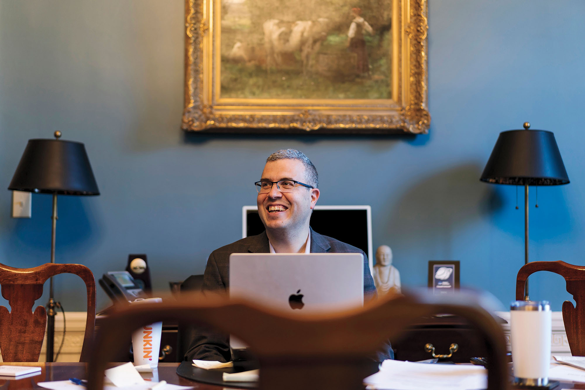 A man wearing glasses and suit sits in a well-appointed office at a desk, behind a laptop computer and smiles.