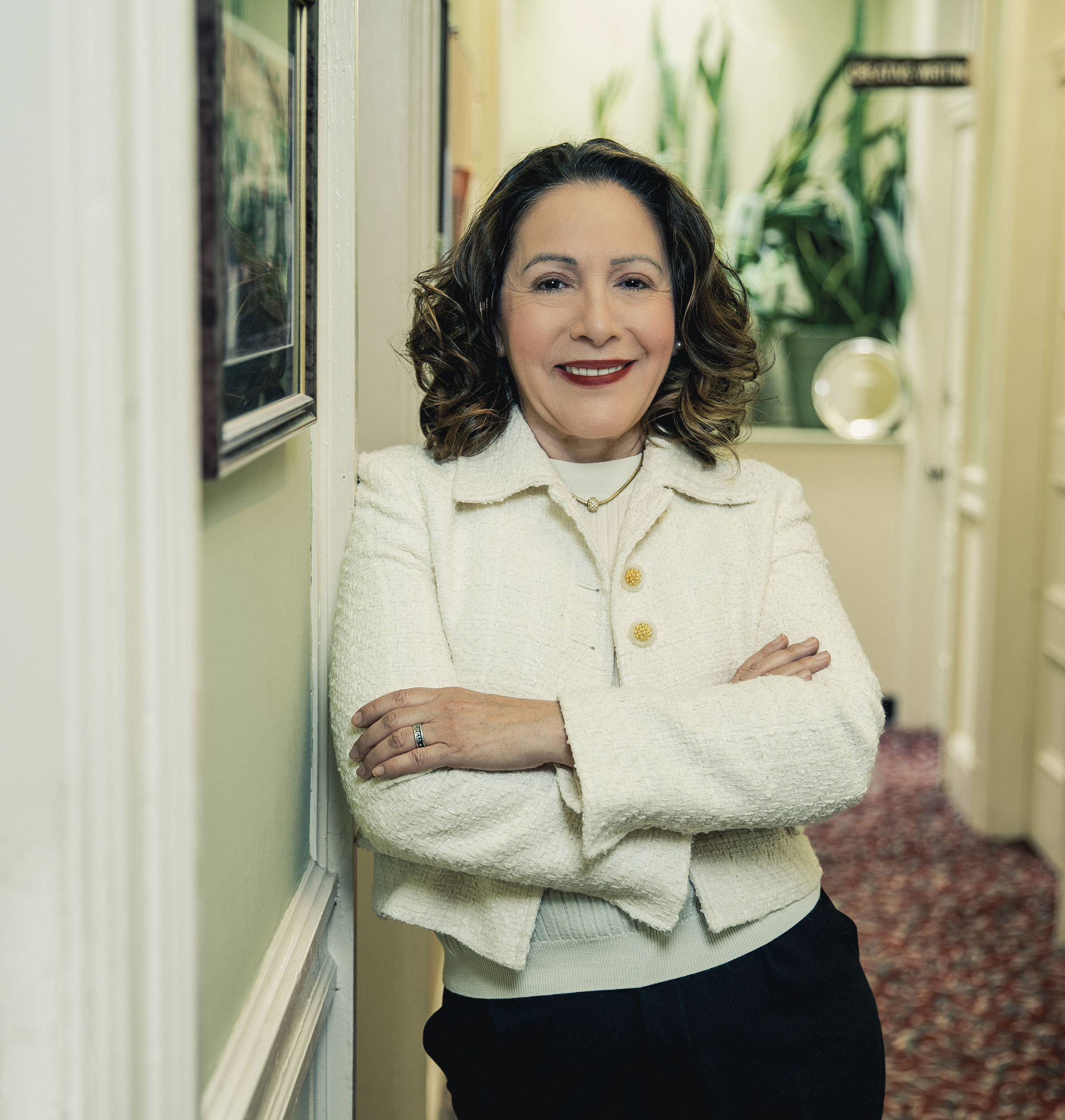 A woman leans against the hallway near academic offices. She wears bright red lipstick, a blazer and slacks, and her arms are folded across her chest.