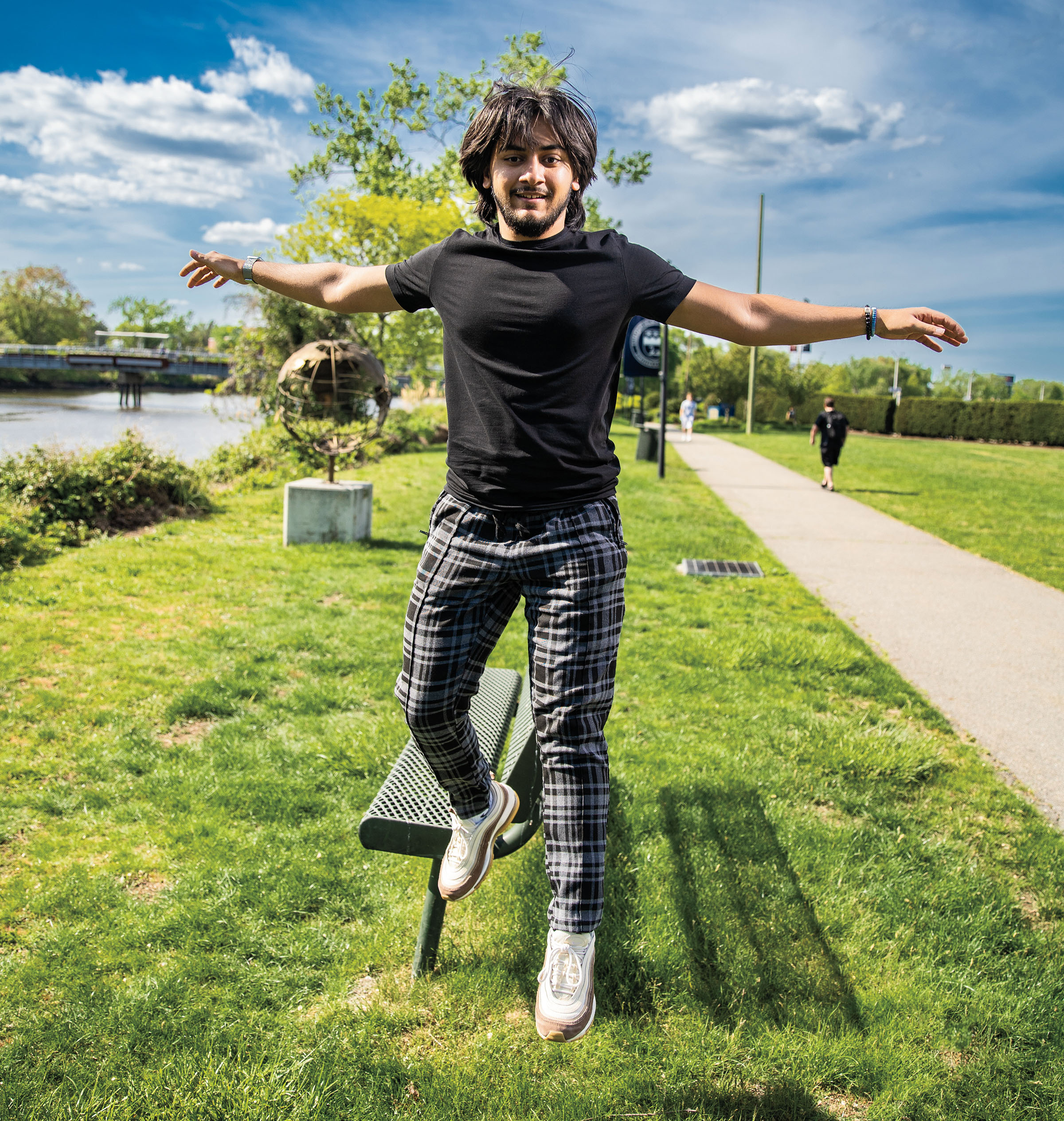 A young man photographed in mid-air, jumping off an outdoor bench in the grass on campus.