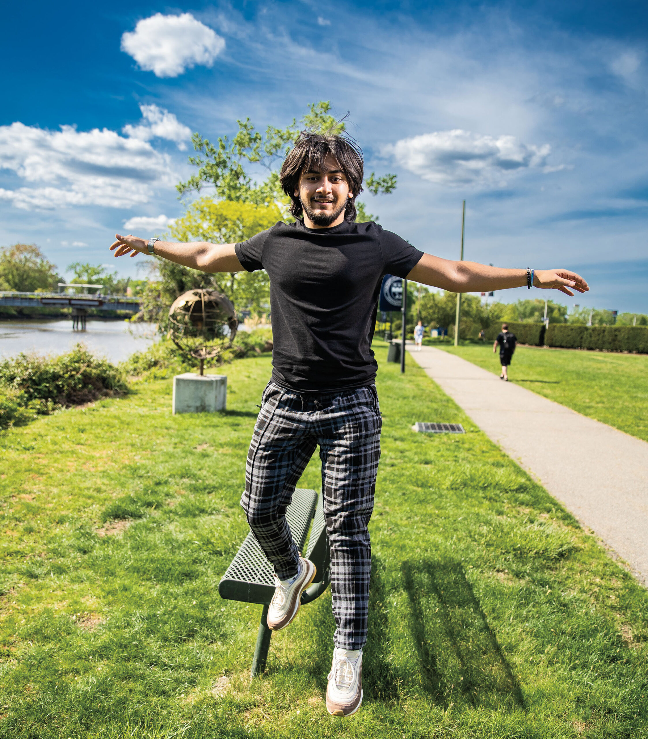 A young man photographed in mid-air, jumping off an outdoor bench in the grass on campus.