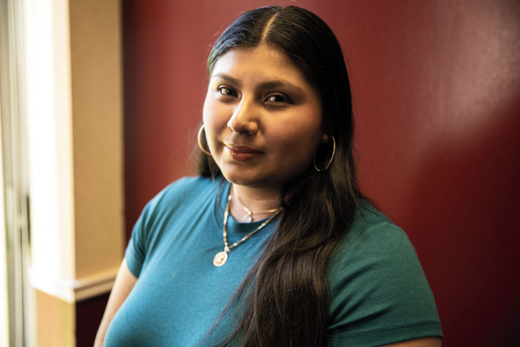 Portrait of a young woman with long dark hair. She stands in front of a red wall.