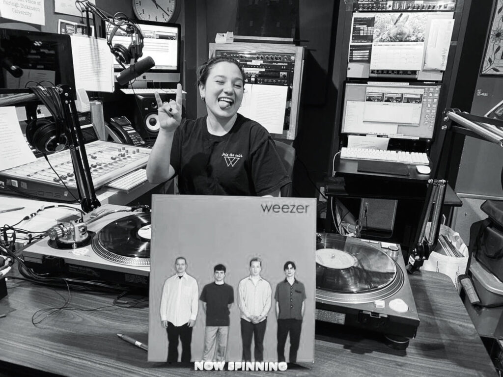 A photo in black and white shows a young woman in a DJ booth at a radio station. She is spinning a vinyl album.