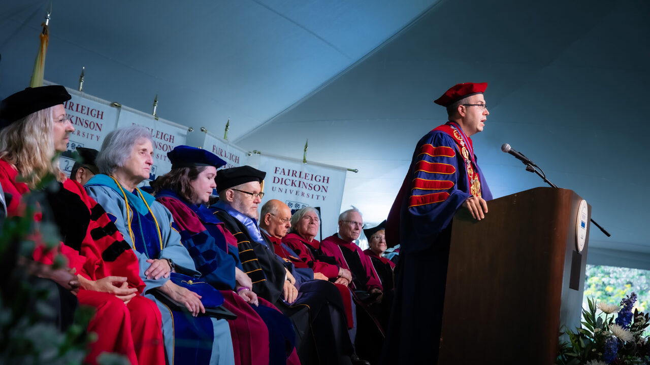 A man in academic regalia and wearing glasses speaks at a podium.