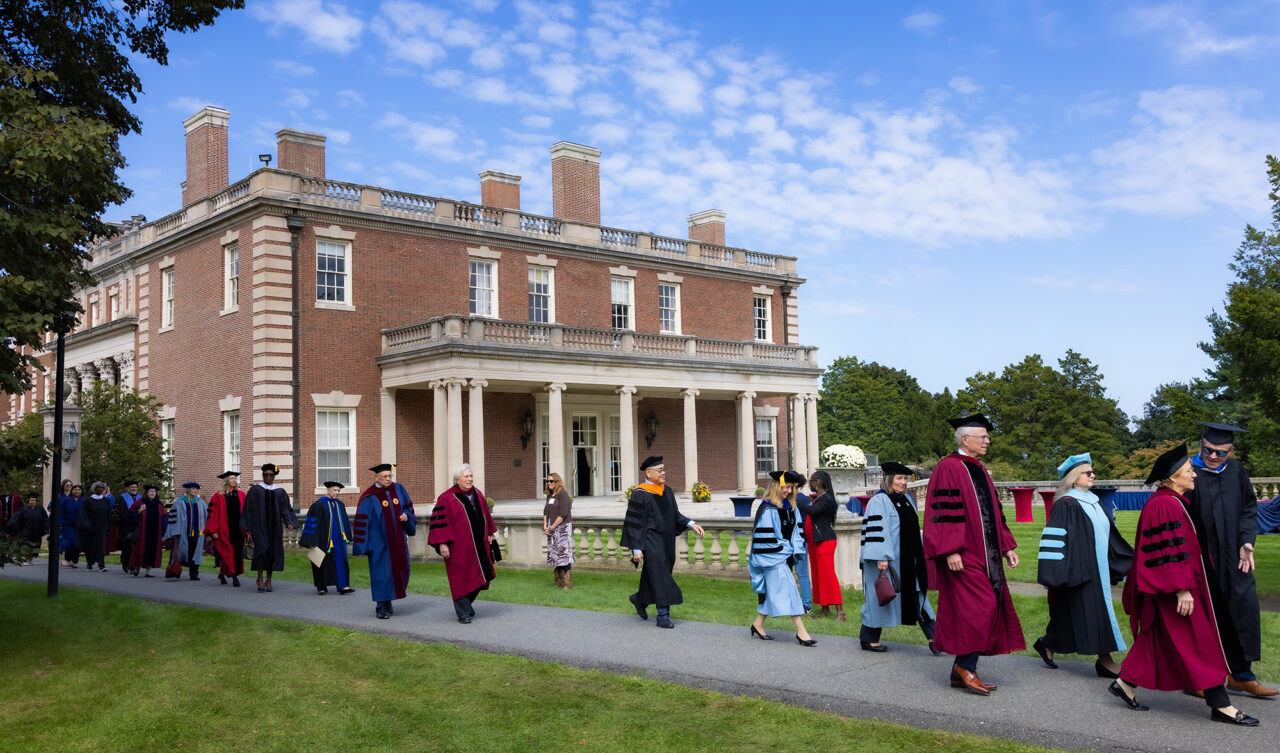 Faculty in academic regalia walk outside.