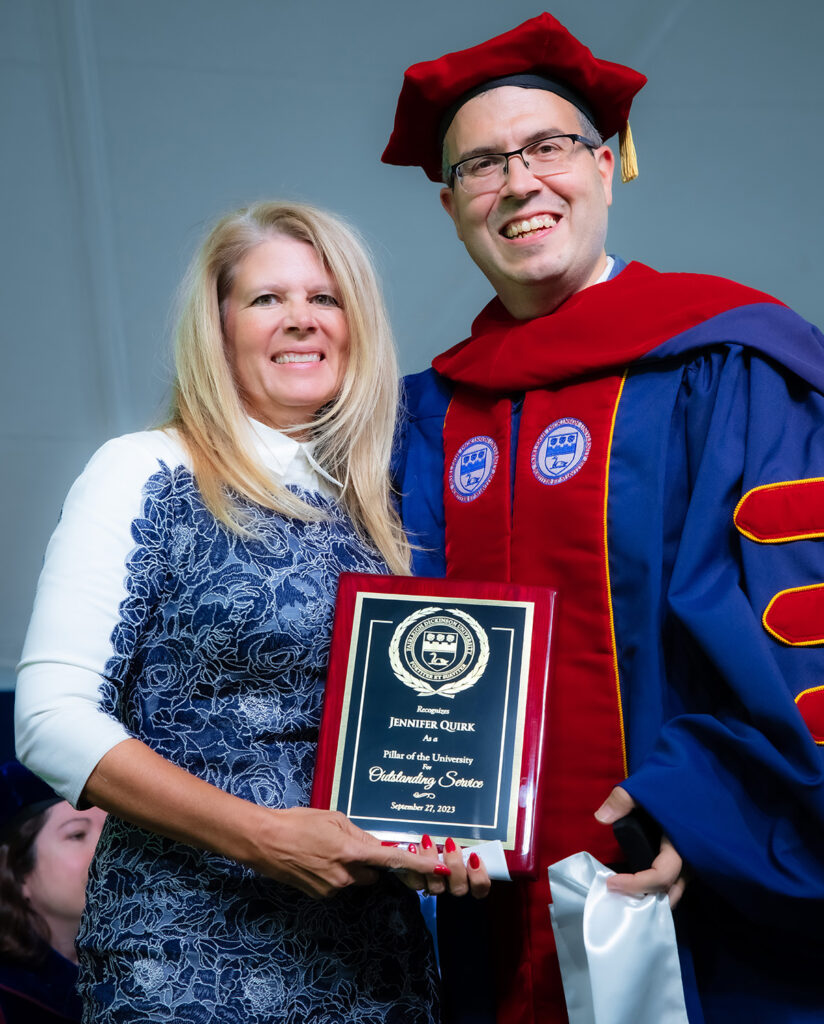 A tall blonde woman holds up an award and stands next to a man in academic regalia.
