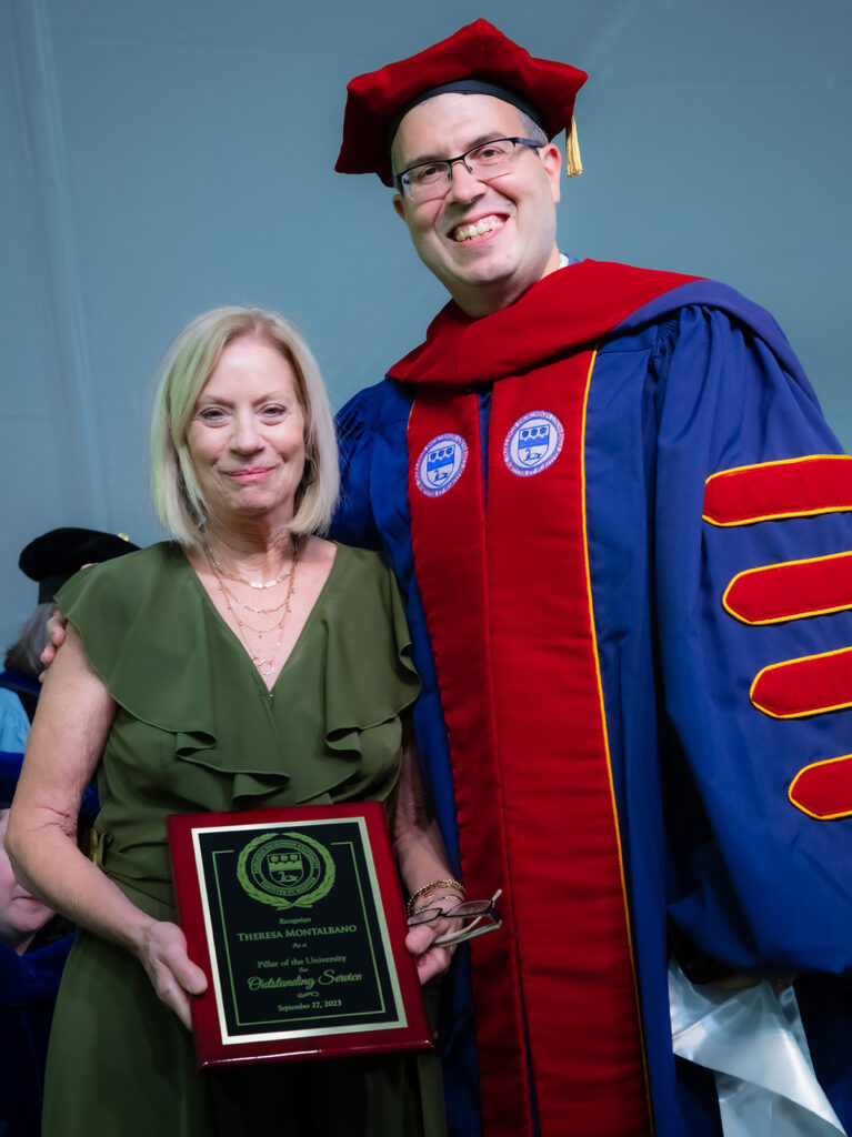 A short blonde woman holds up an award and stands next to a man in academic regalia.