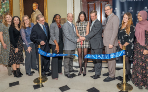 A group of men and women gather to cut a ribbon and officially dedicate a new office on campus.