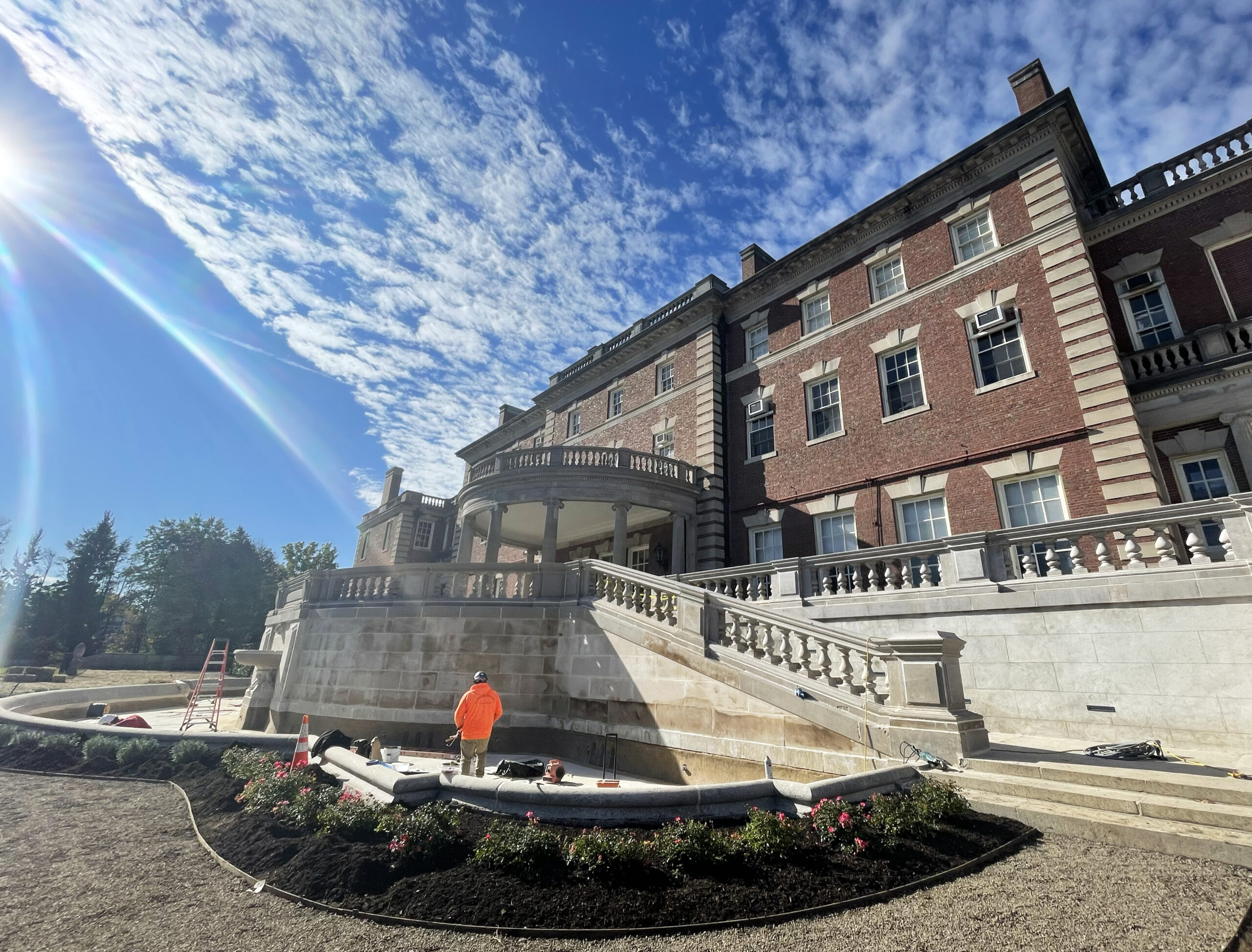 The back of a stately, historic mansion, with a restored terrace and portico, fountain and new landscaping. A man in construction gear surveys the scene.