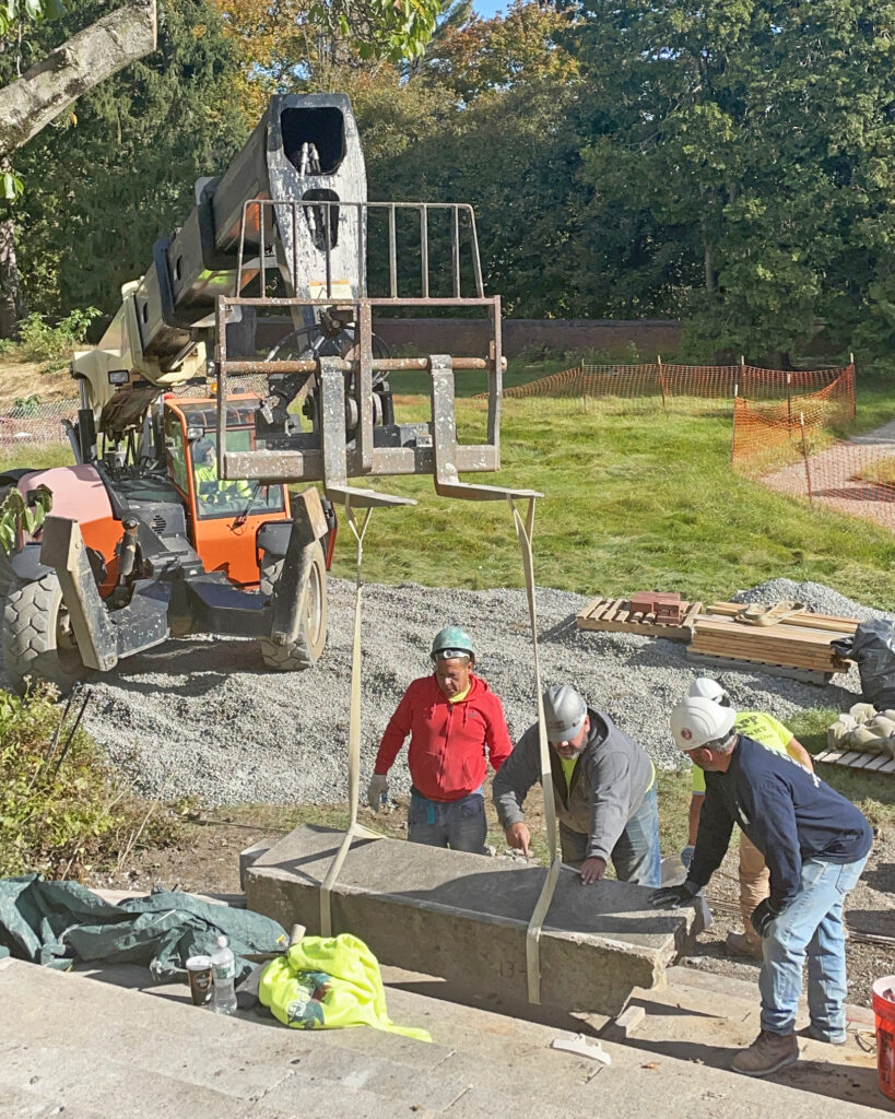 Workers use construction vehicles and equipment to raise a large stone step.