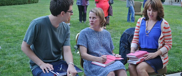 Three people sitting on a lawn, holding books and talking