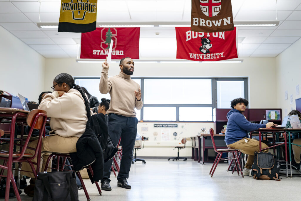 An African American man in a tan sweater teaches students in a classroom. 