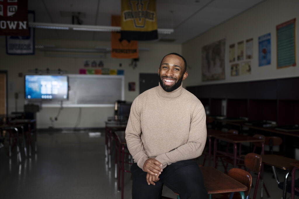 An African American man in a tan sweater sits in a classroom smiling for a portrait. 