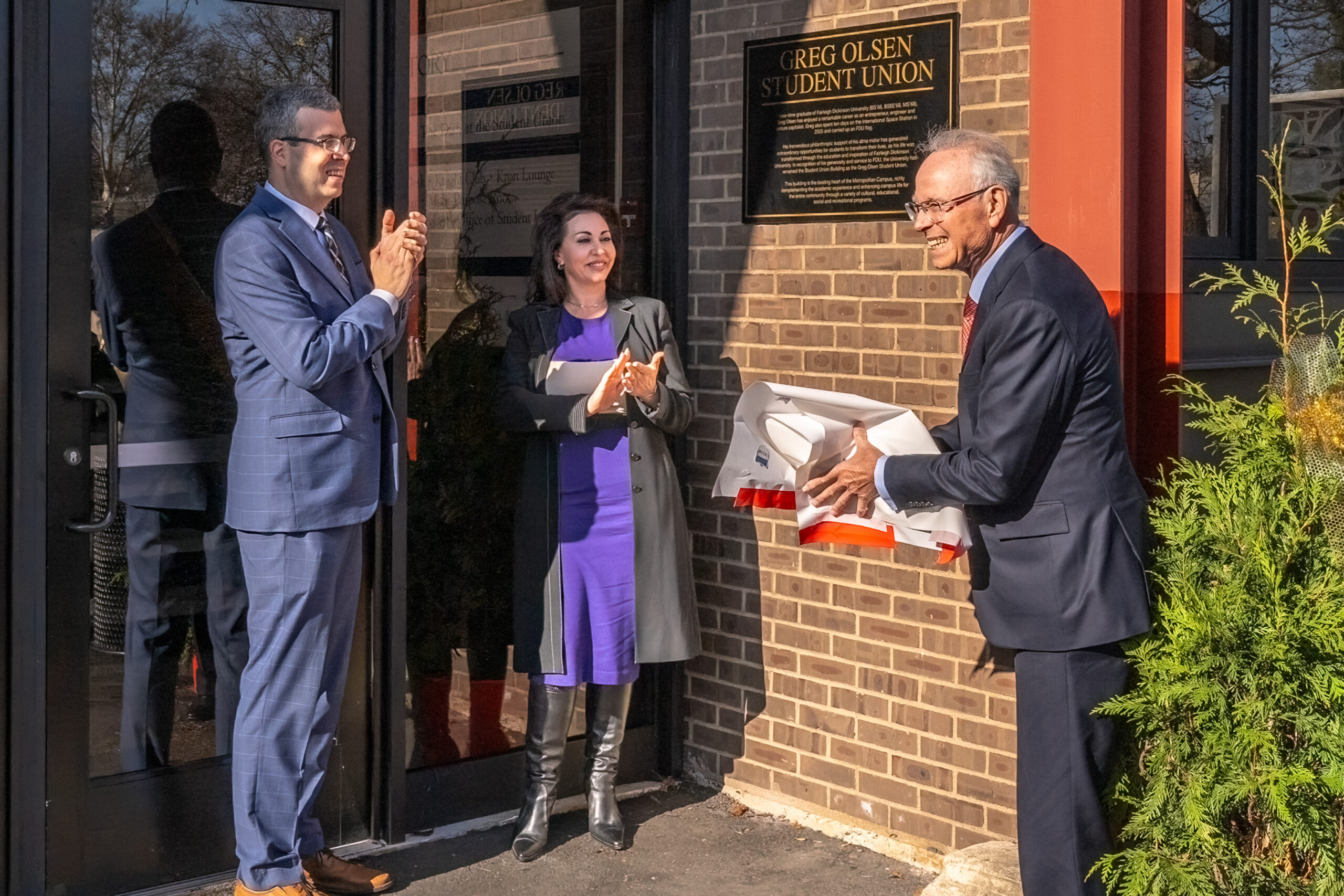 A man in a blue suit claps next to a woman in a purple dress, while a man holds a white paper next to a sign and building. 