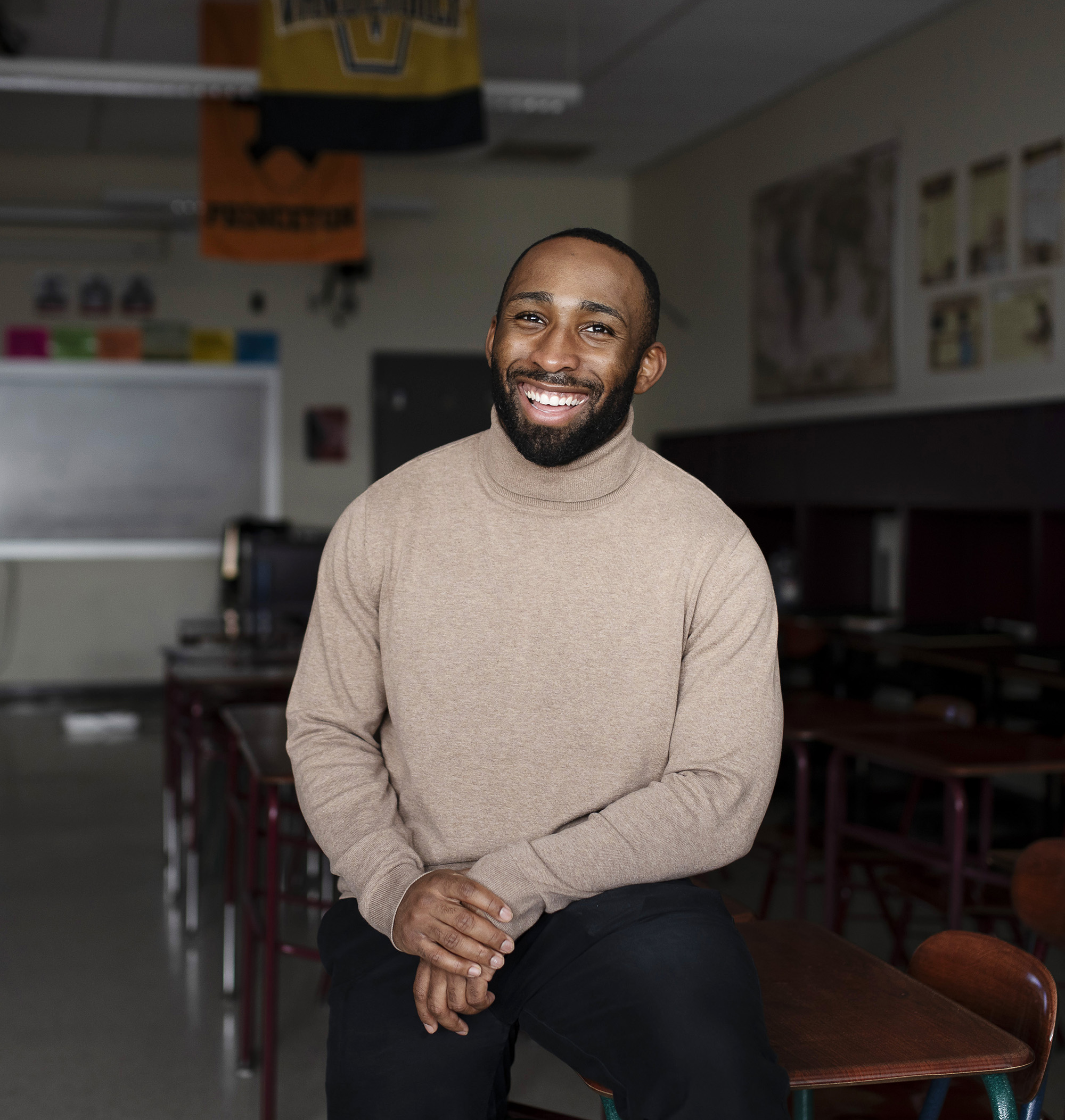 A young man in tan sweater sits on a desk in a darkened classroom.