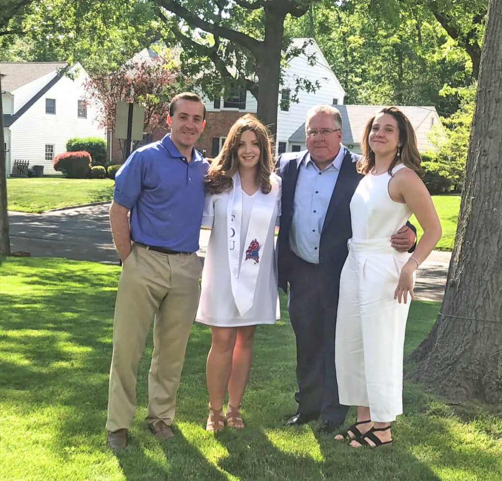 A man in blue stands next to a woman in a white dress. Next to her a man in a black suit with a blue shirt stands next to a woman in a white jumpsuit. They are outside on green grass. 