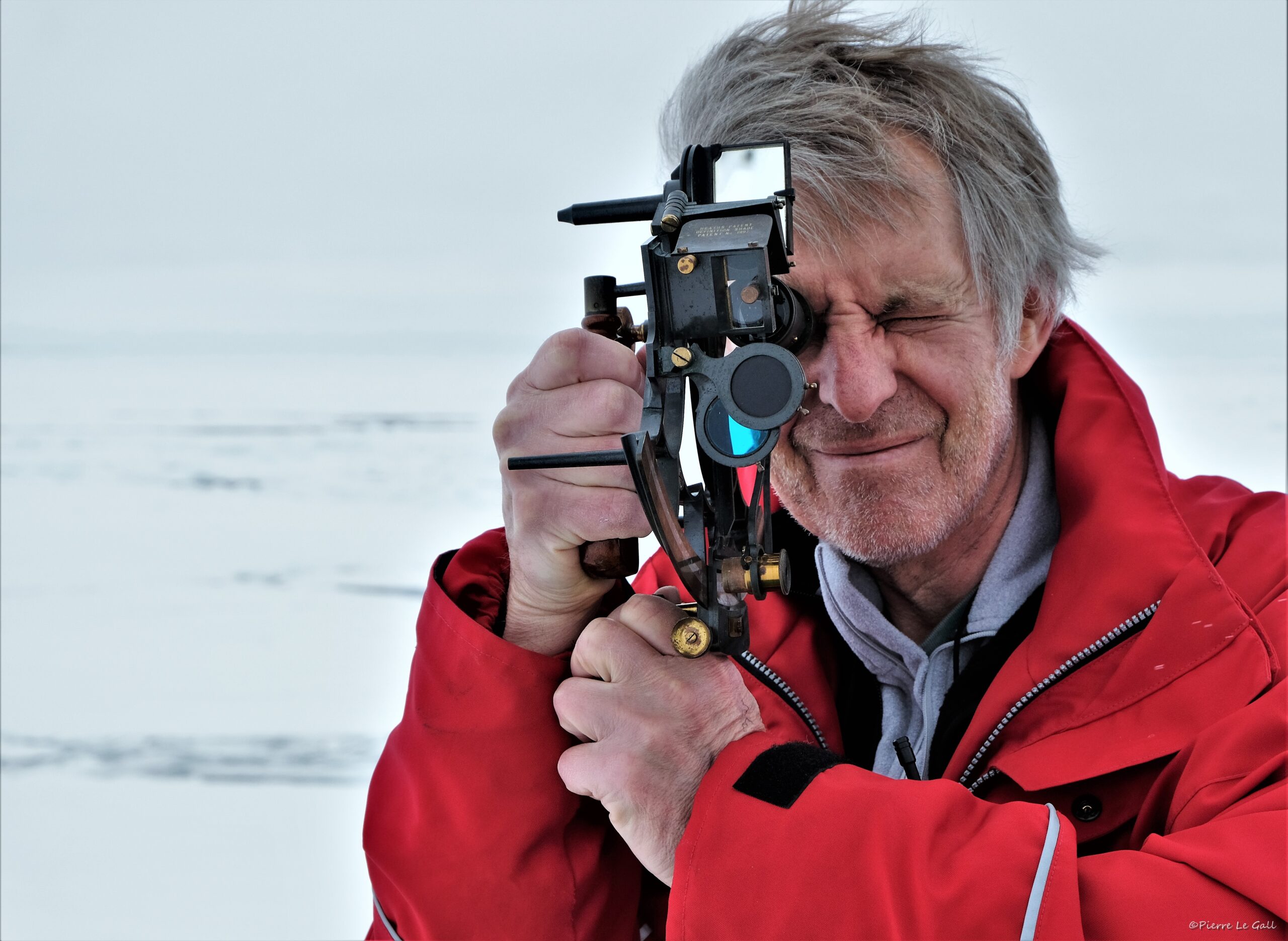 A man wearing a red parka coat takes an angle on the sun with a sextant.
