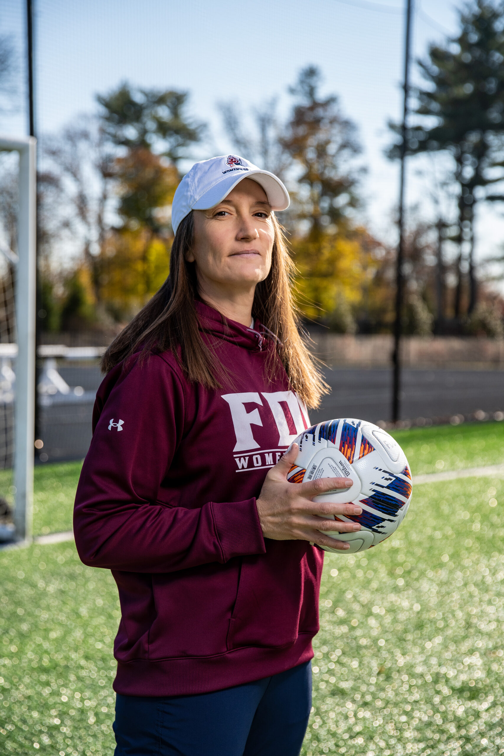 A woman in a white baseball cap and a burgundy shirt stands on a soccer field holding a soccer ball. 