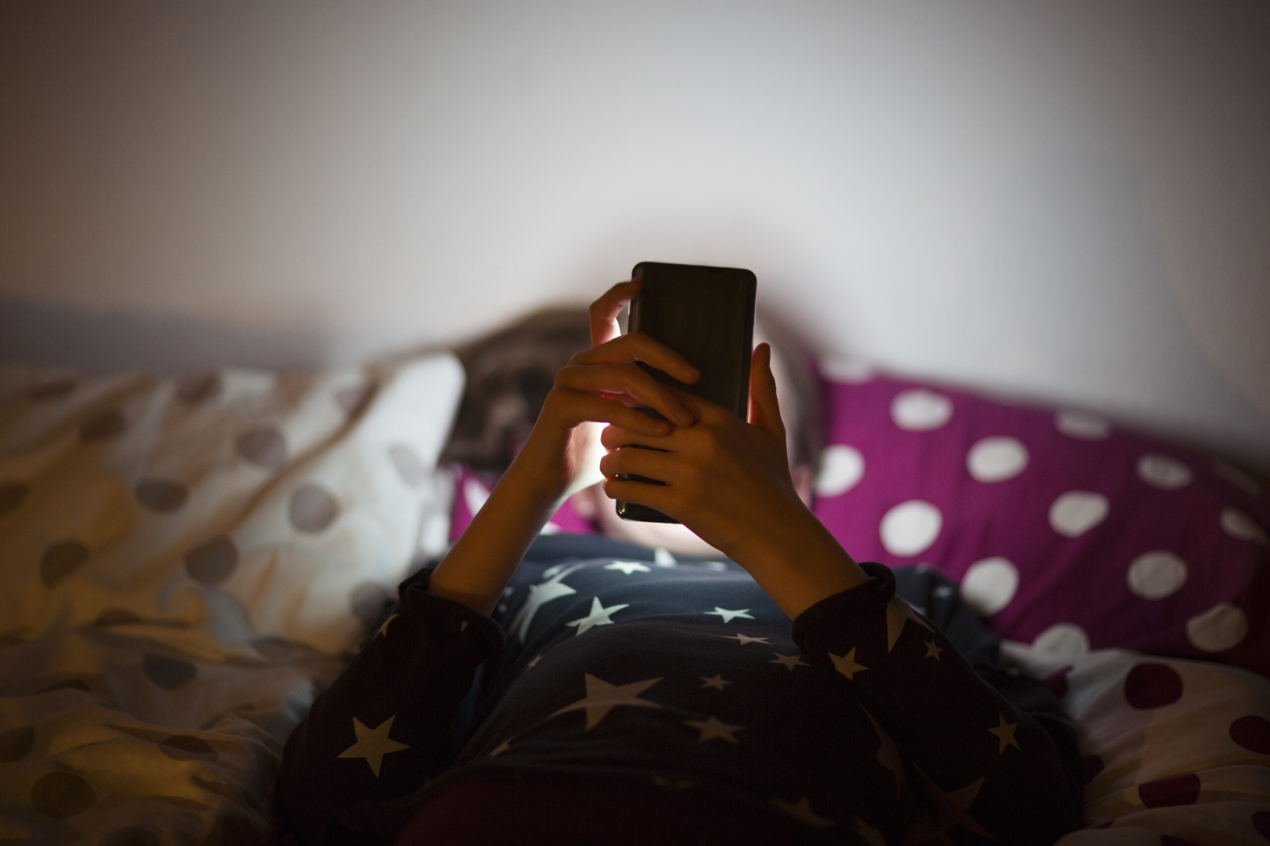 A young woman relaxes on a bed, and scrolls through her smartphone.