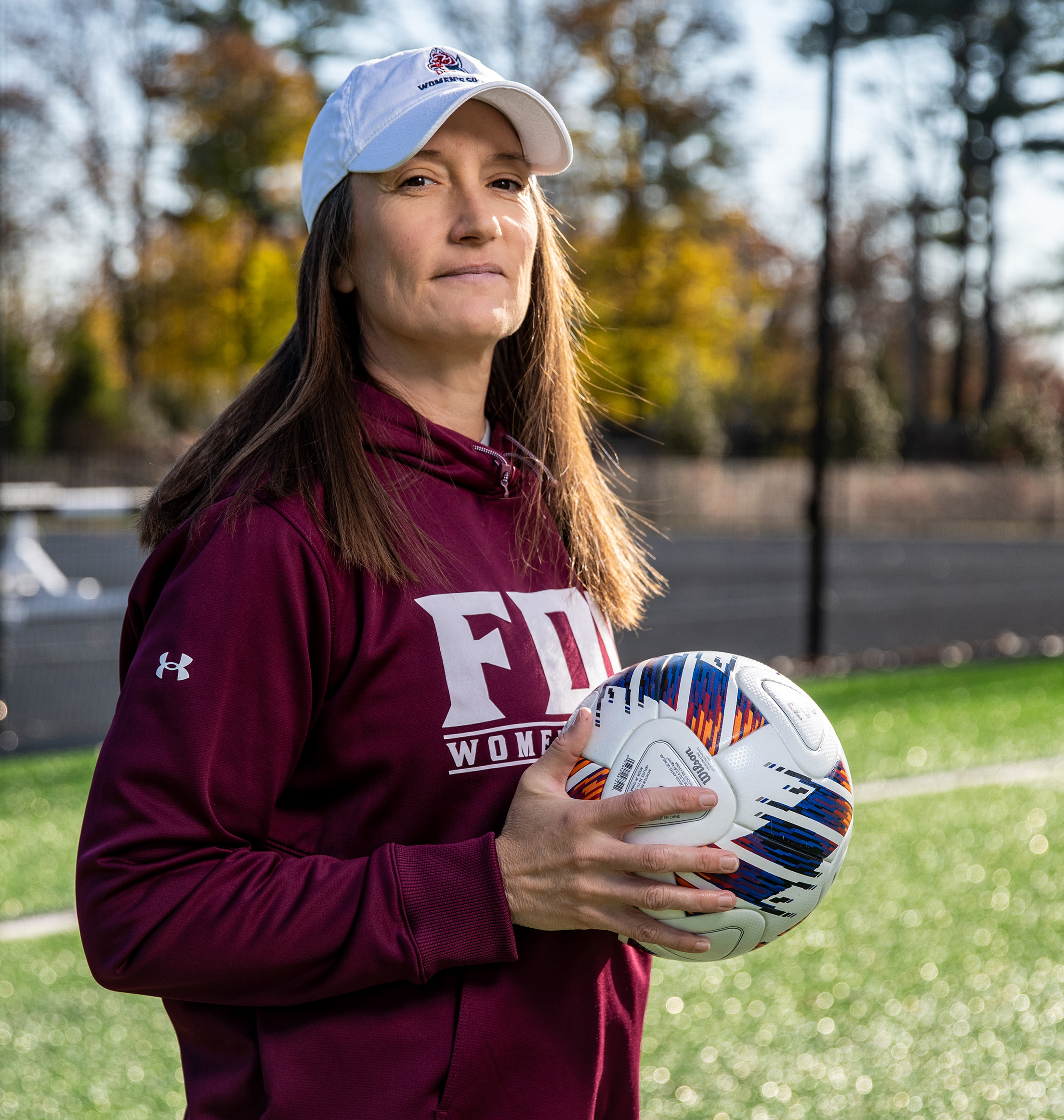 A woman in a white baseball cap and a burgundy shirt stands on a soccer field holding a soccer ball.