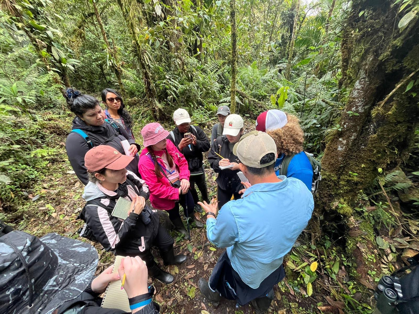 a group of students huddle around a professor. they are standing in a rainforest. they hold notebooks and observe a insect held by the professor.