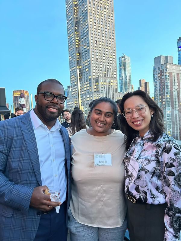 An African American man in a blue suit stands with an Indian woman in a beige shirt and an Asian women in a floral pink shirt. The New York City skyline is behind them.  