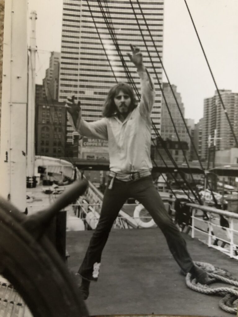 A vintage black and white photos shows a young man with long hair and facial hair, jumping at the South Street Seaport Museum in Manhattan.