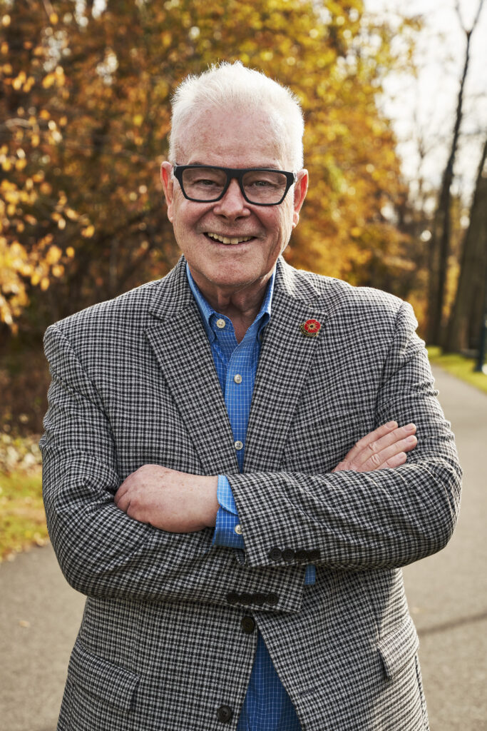 A man wearing glasses and a tweed jacket poses for a photo, arms crossed.