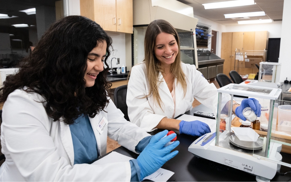 two students in white lab coats smile as they measure medicine.