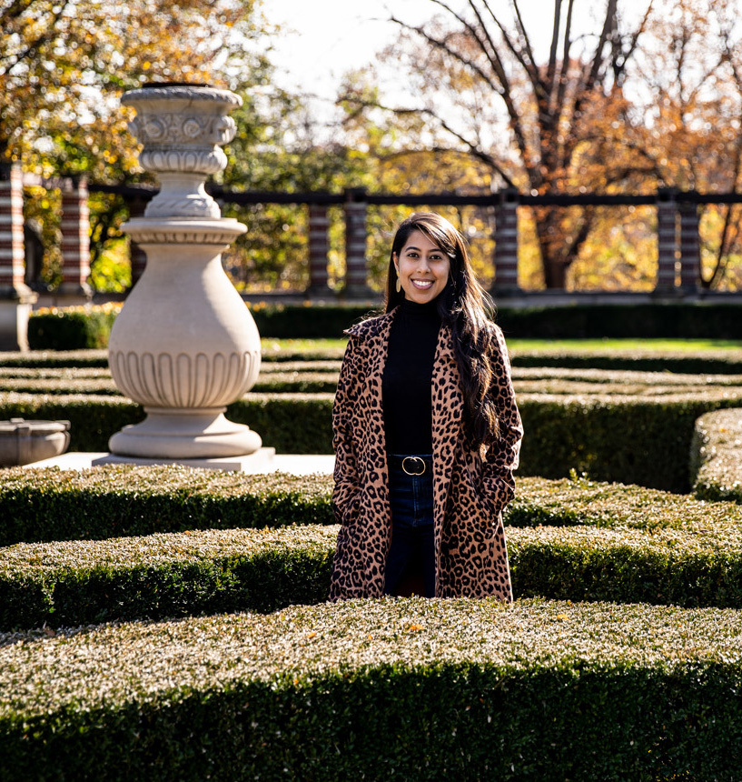A young woman wearing a leopard print coat, stands in the middle of a garden and smiles.