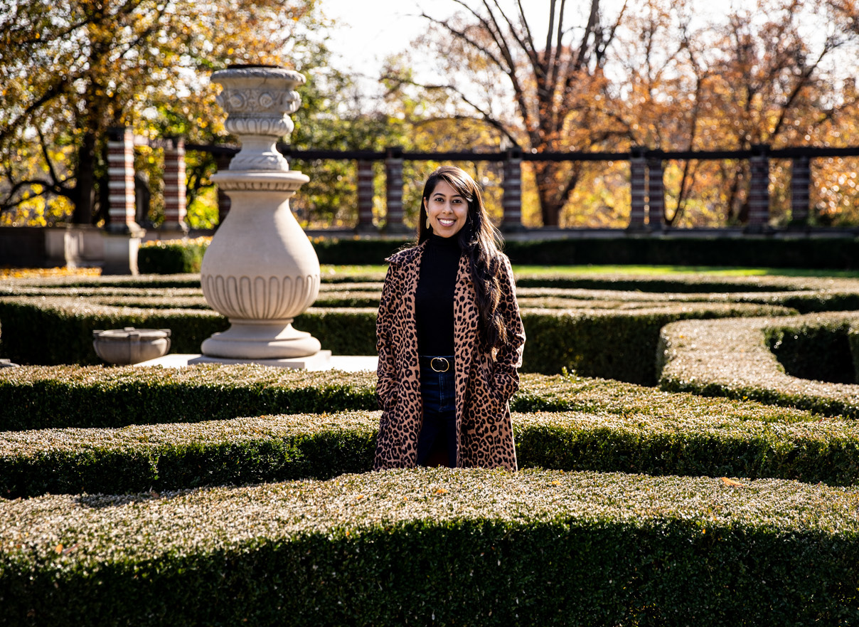A young woman wearing a leopard print coat, stands in the middle of a garden and smiles.