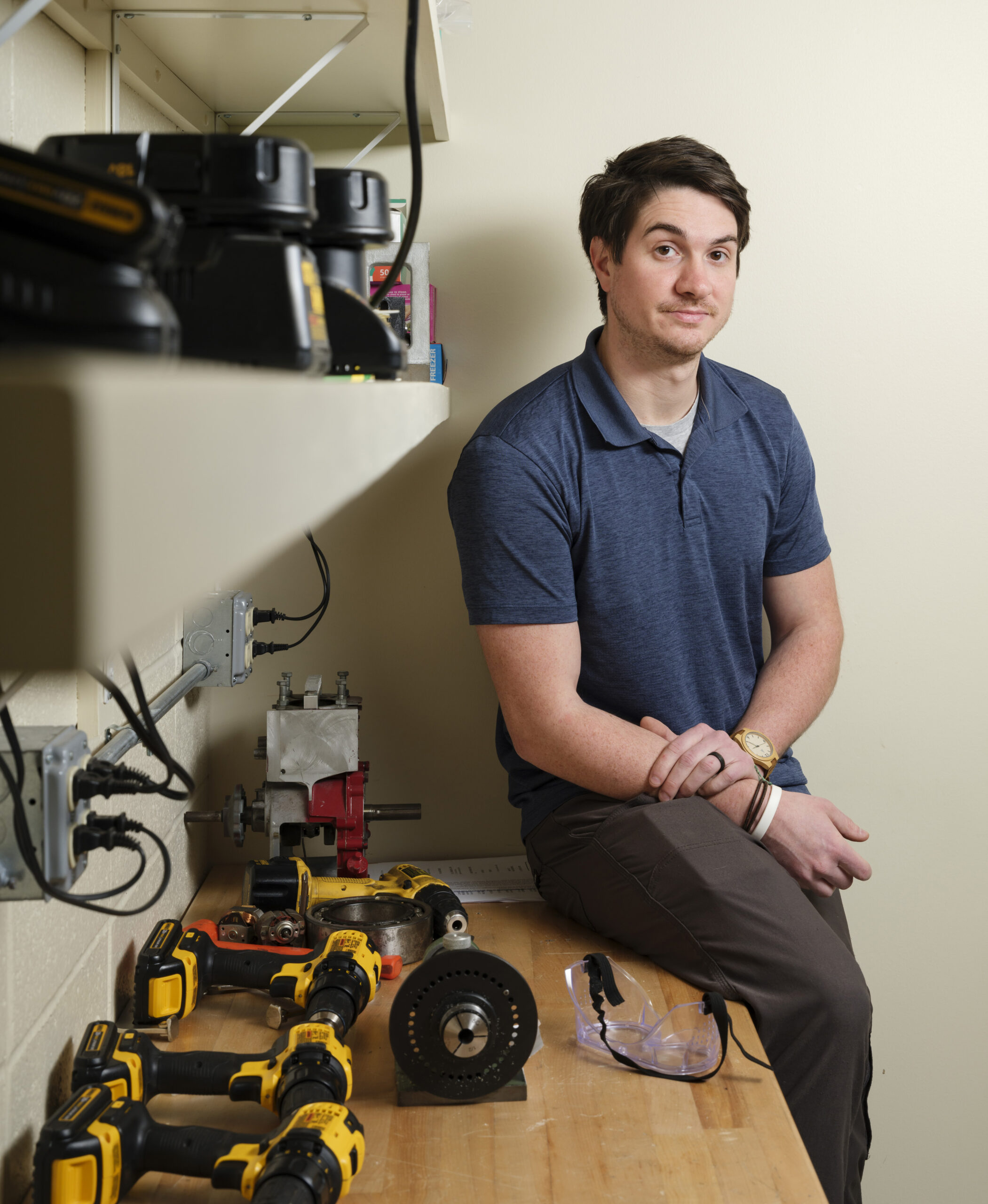 A man sits on the edge of a table next to various electronic devices and tools.