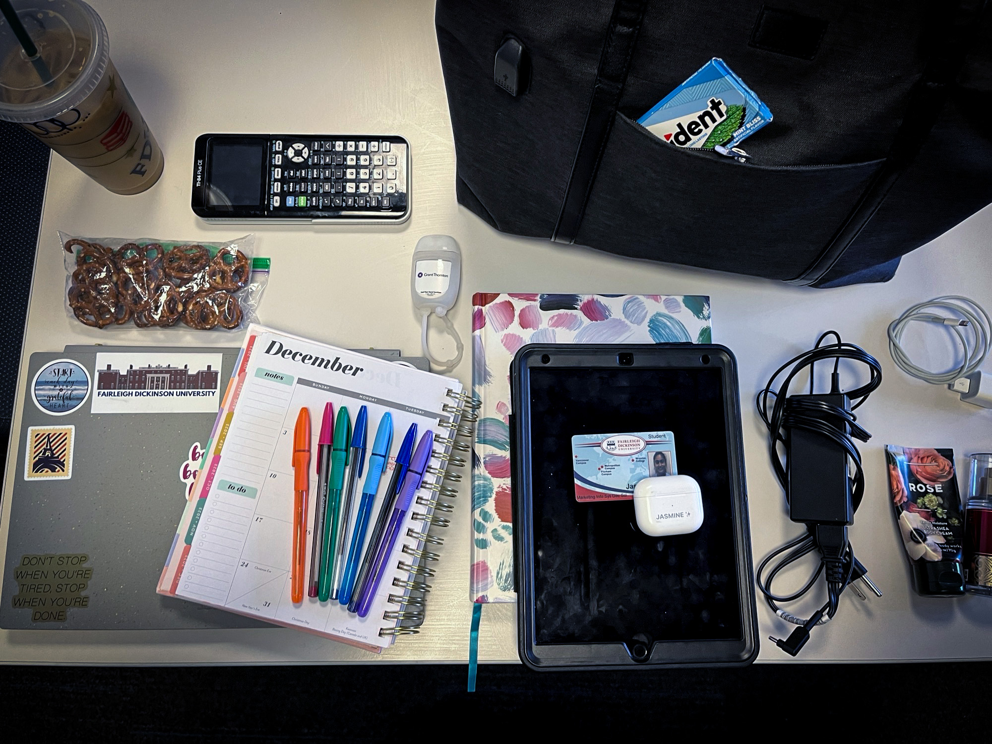 Colorful pens, a University ID, cords and chargers, a laptop, snacks, a calculator and a pack of gum sit atop a desk.
