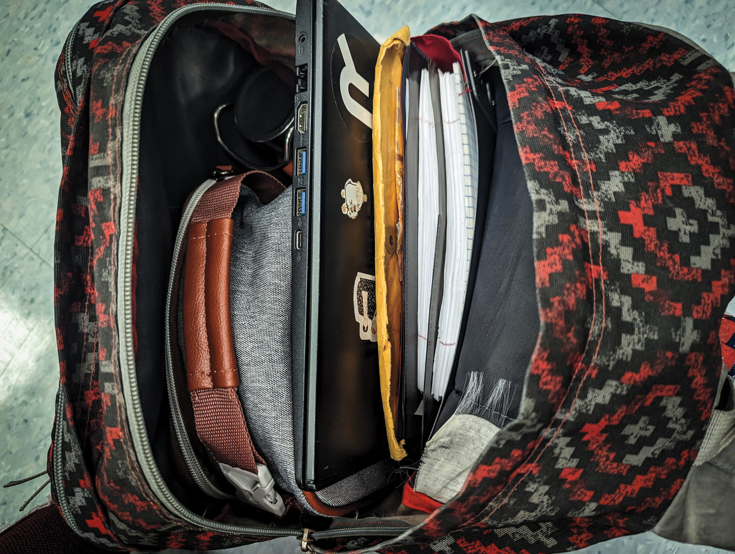 An open red and gray patterned backpack, with a lunchbox, laptop and notebooks visible.