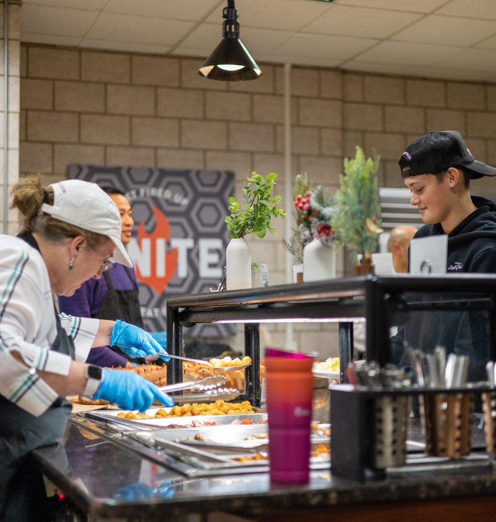 a student being served food in the dining hall. the employee scoops tater tots.