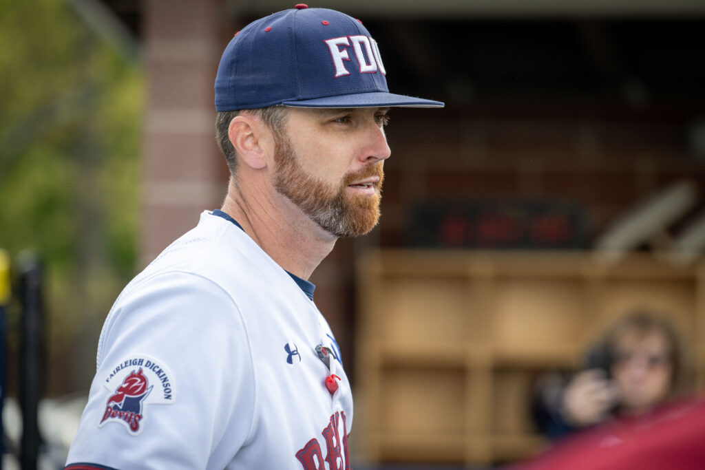 A man with facial hair stands on a baseball field wearing a hat and uniform. 