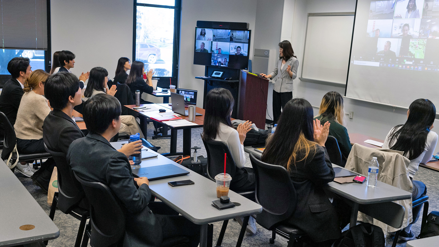 An instructor stands at the front of the class and teaches a room of students in person and on Zoom.