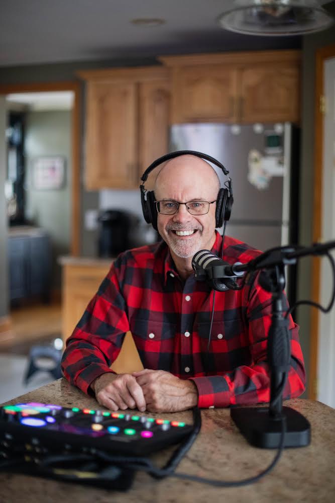 A man sits at a kitchen table. He is wearing headphones and had audio podcast equipment in front of him. 