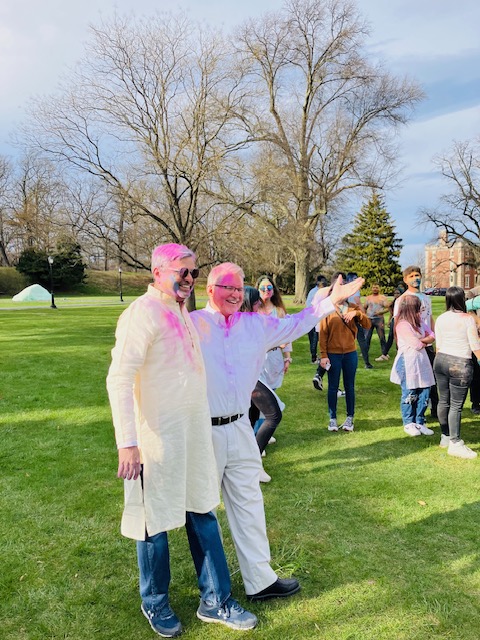 Two men stand together in white clothing covered in pink powder to celebrate Holi. 