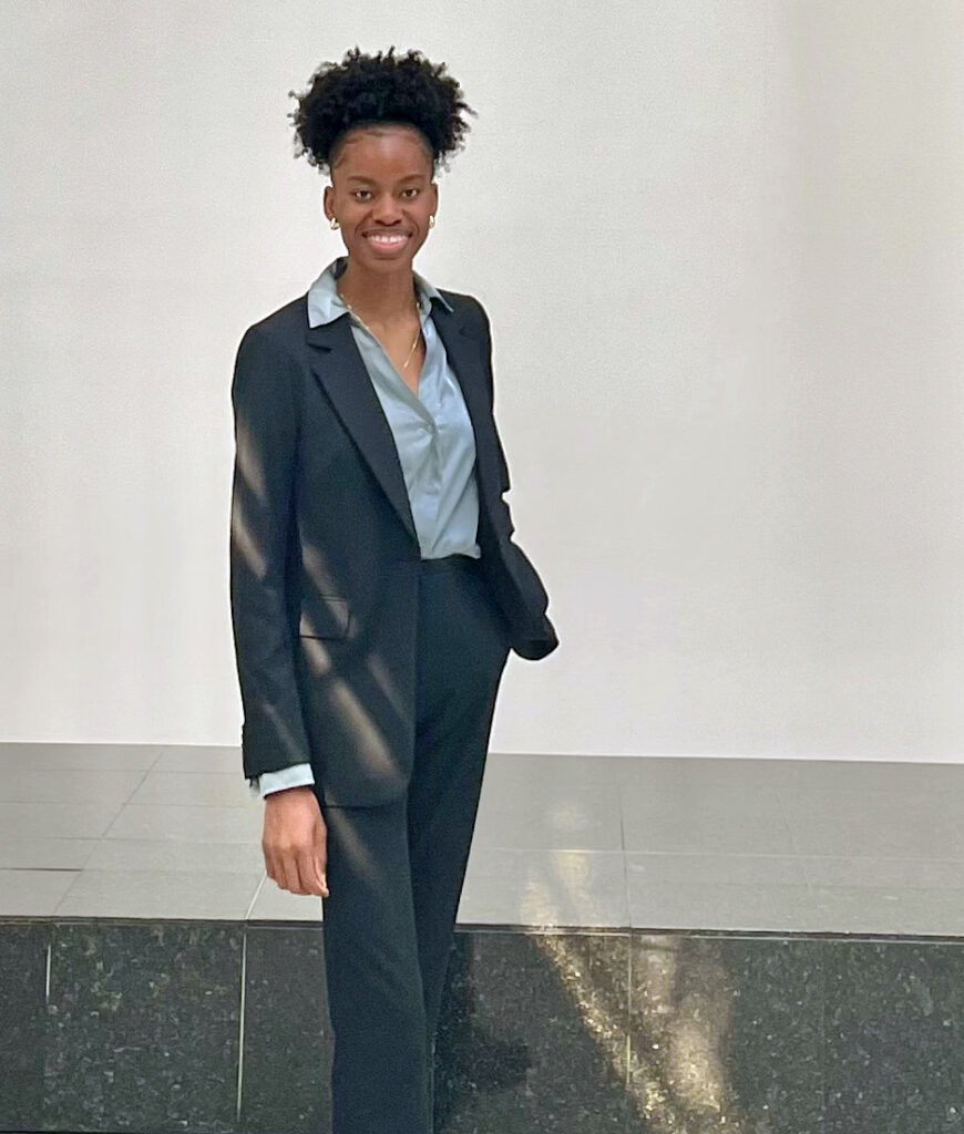 A young woman wearing a pantsuit poses standing and smiles for a photo.